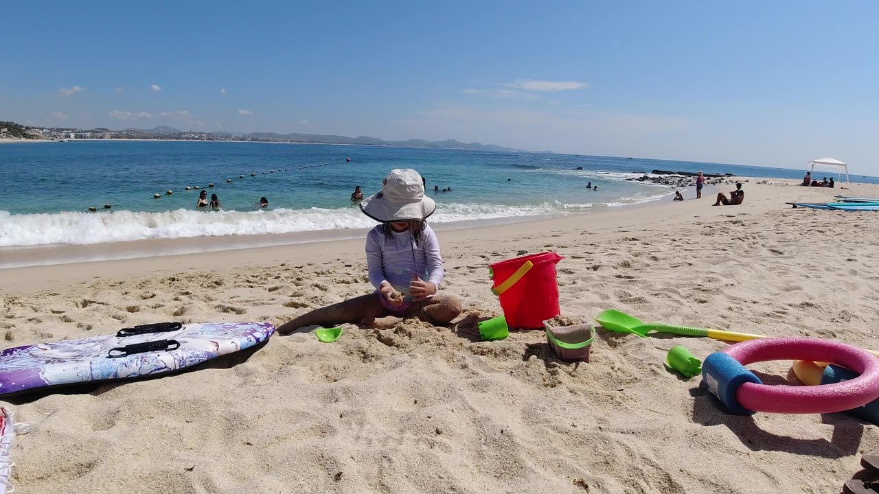 The child remains seated on the Los Cabos beach, looking down at the sand while a red bucket, green sand toys, bodyboard, inflatable ring, swimmers, and a white canopy sit around the shoreline.