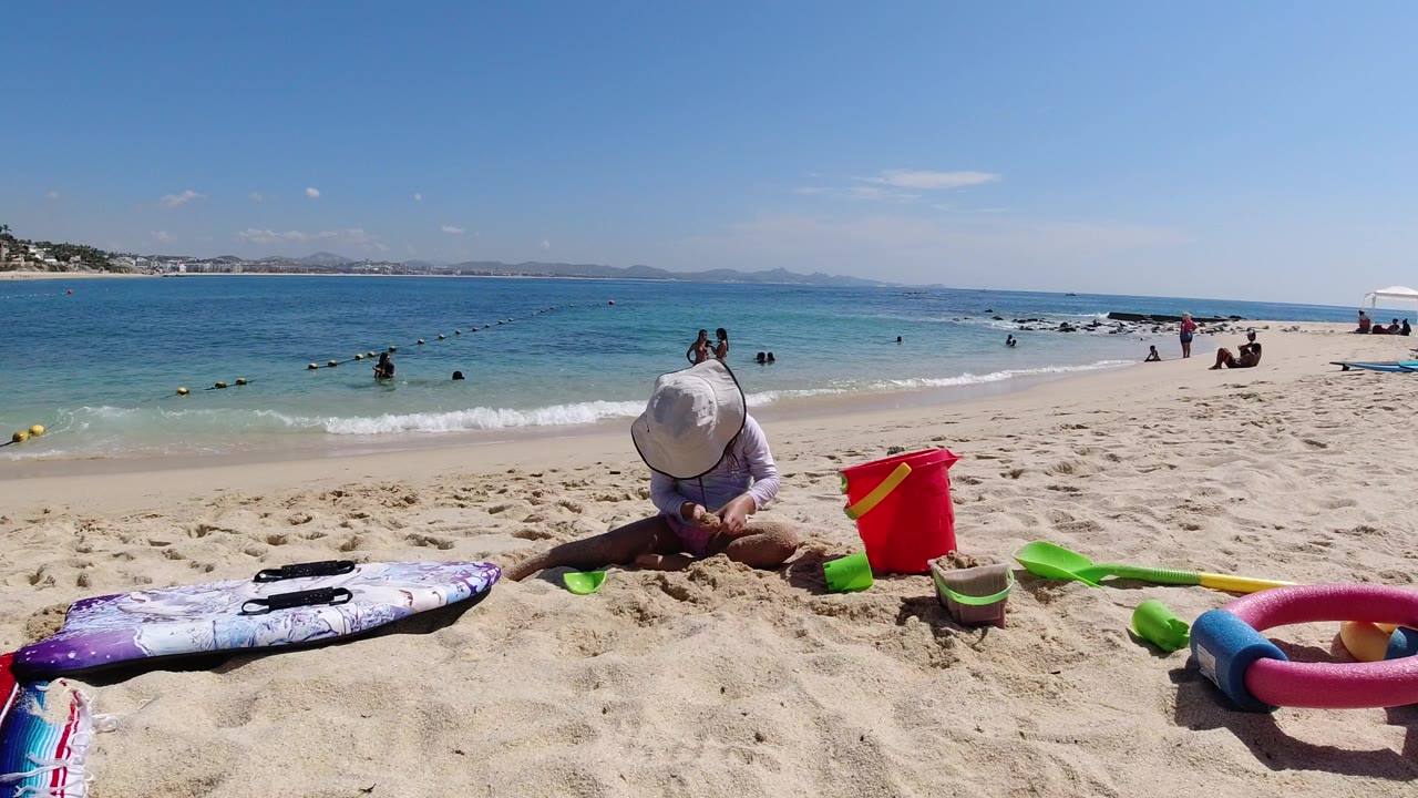 Child in sun hat playing in sand beside beach toys and turquoise water.
