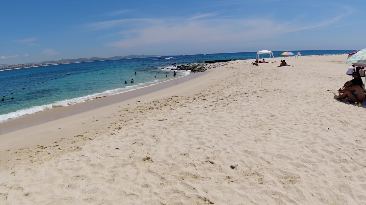 Wide sandy beach with blue water, umbrellas, and swimmers offshore.