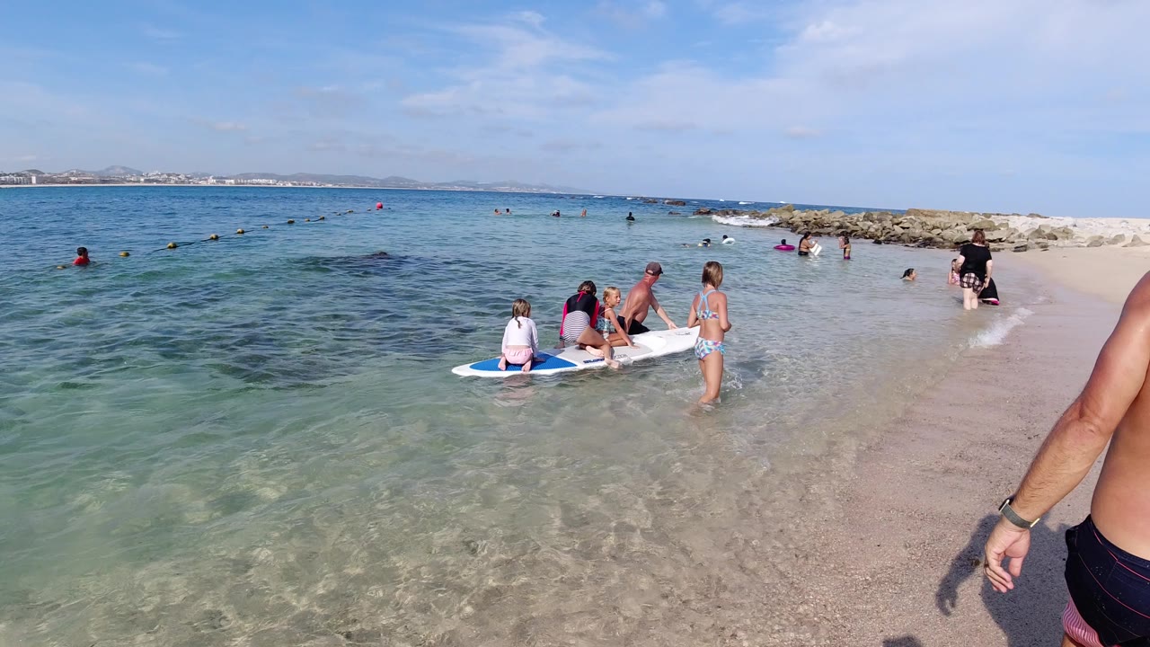 Children gather on and beside a white paddleboard in shallow water at a Los Cabos beach, with adults supervising nearby and swimmers beyond the rocky breakwater.