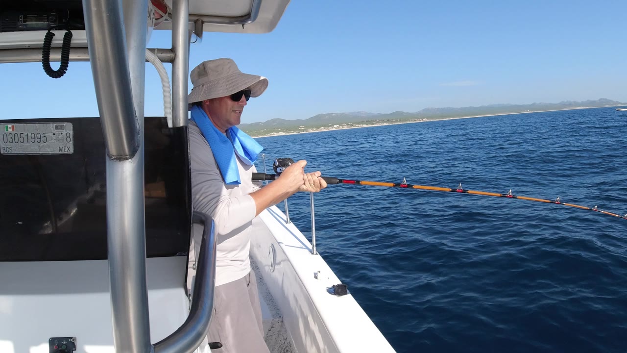 At 0:01, the angler remains near the boat canopy support while the fishing rod arcs toward the water and the Los Cabos coastline runs along the horizon.