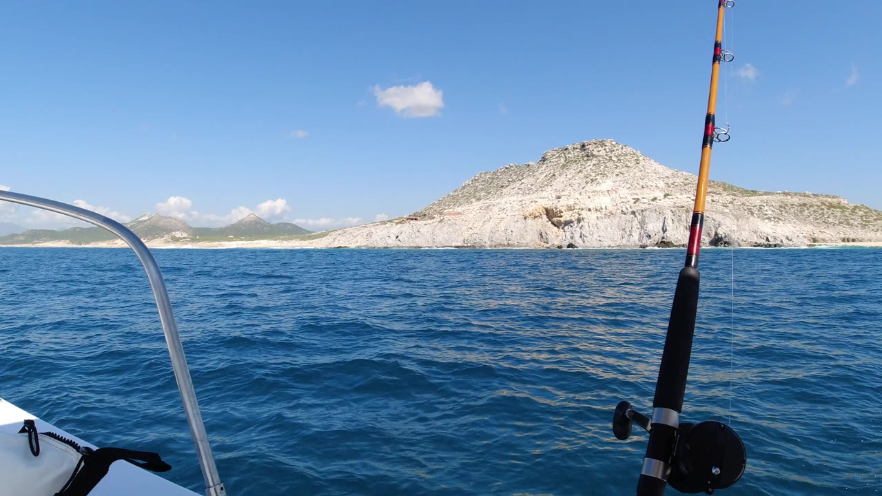 Opening frame from Los Cabos, Mexico showing a fishing rod at right, a curved boat rail at left, blue rippled water, a pale rocky hill, and no visible people.