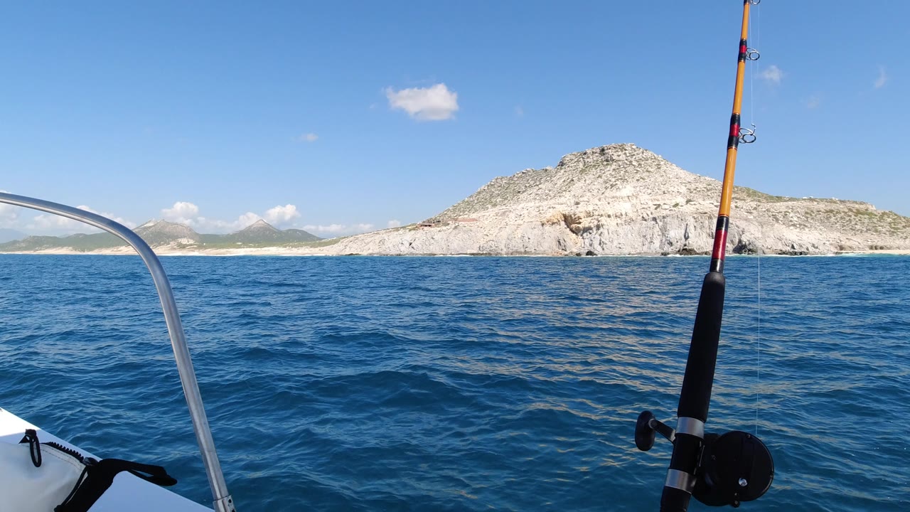 Los Cabos water view with the boat bow edge lower left, metal rail near center, fishing rod on the right, textured blue sea, rocky coastline, and no visible people.