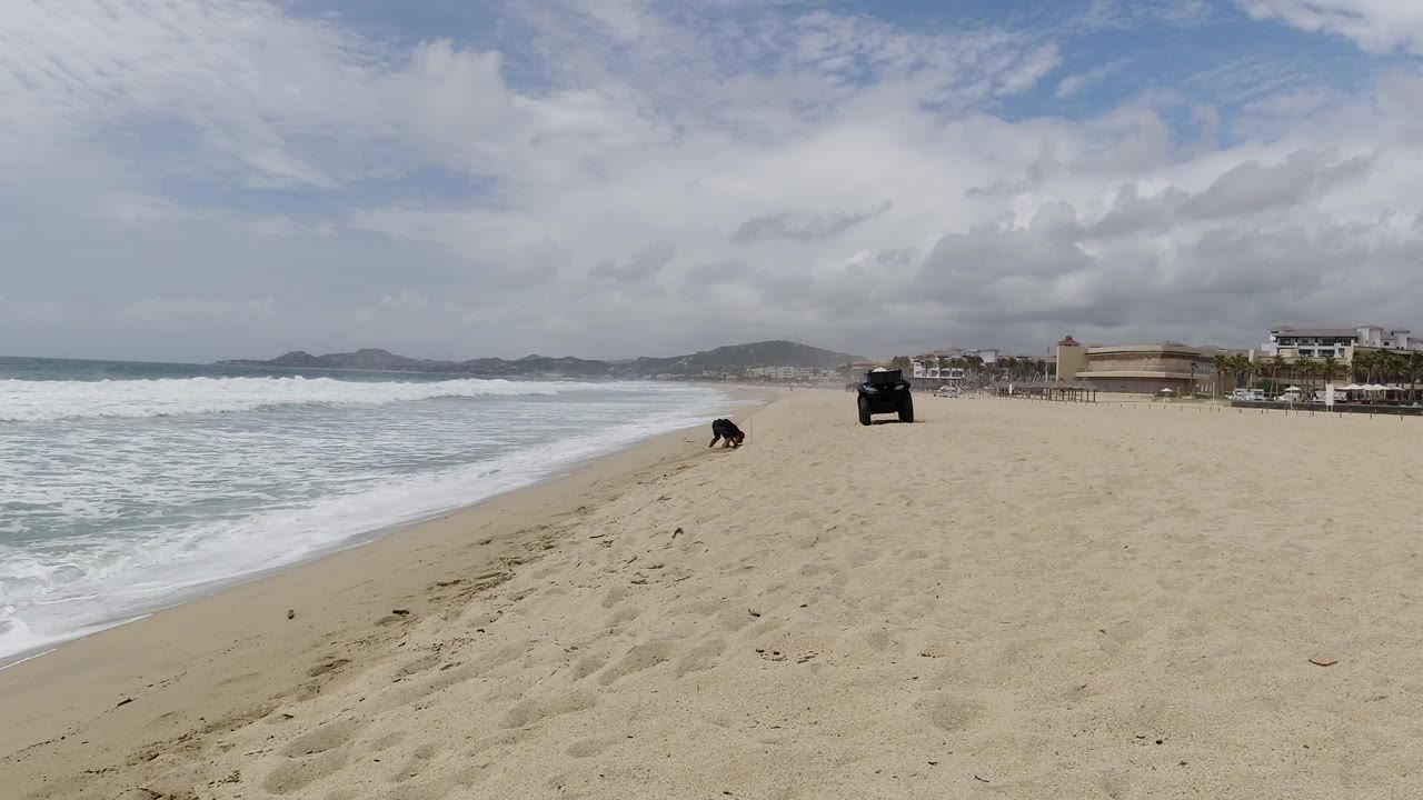 Wide sandy beach with surf, a parked dark vehicle, and a person near the shoreline.