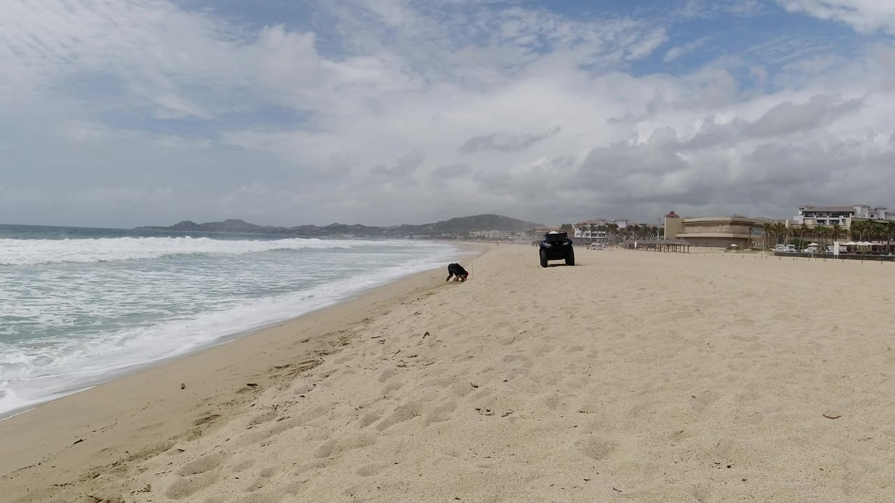 A Los Cabos shoreline view shows the surf edging up the left side, footprints and sand texture in the foreground, the Wrangler farther ahead, and a person near the wet sand.