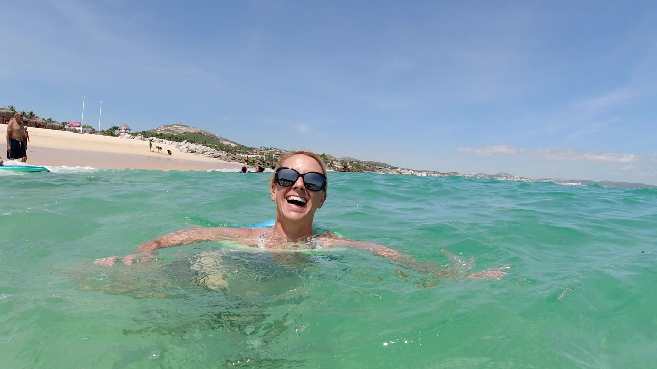 Smiling woman swimming in turquoise water near a sandy beach.