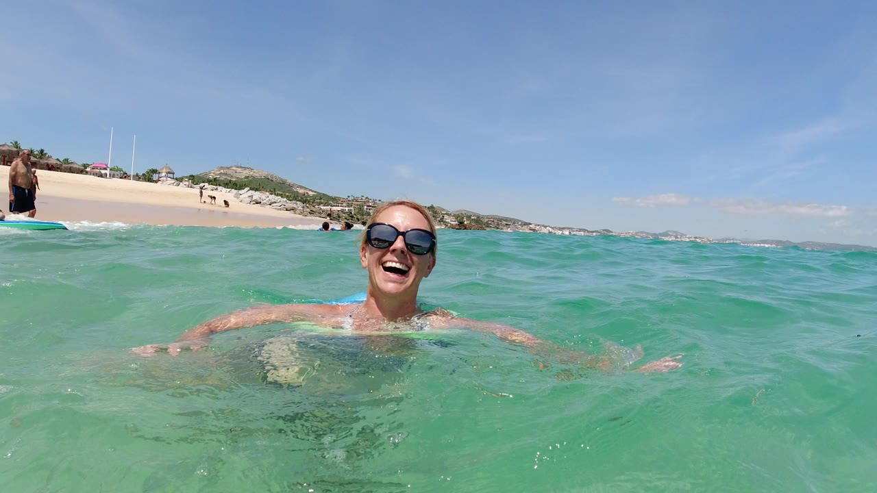 The woman stays centered in the water in Los Cabos, Mexico, smiling toward the camera as small waves cross the foreground and swimmers appear near shore.