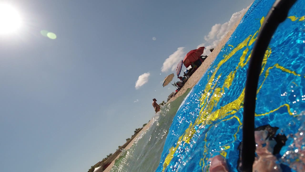 A similar tilted Los Cabos beach view shows the blue board wet with spray in the foreground, a swimmer or beachgoer near the waterline, and umbrellas on the sand.