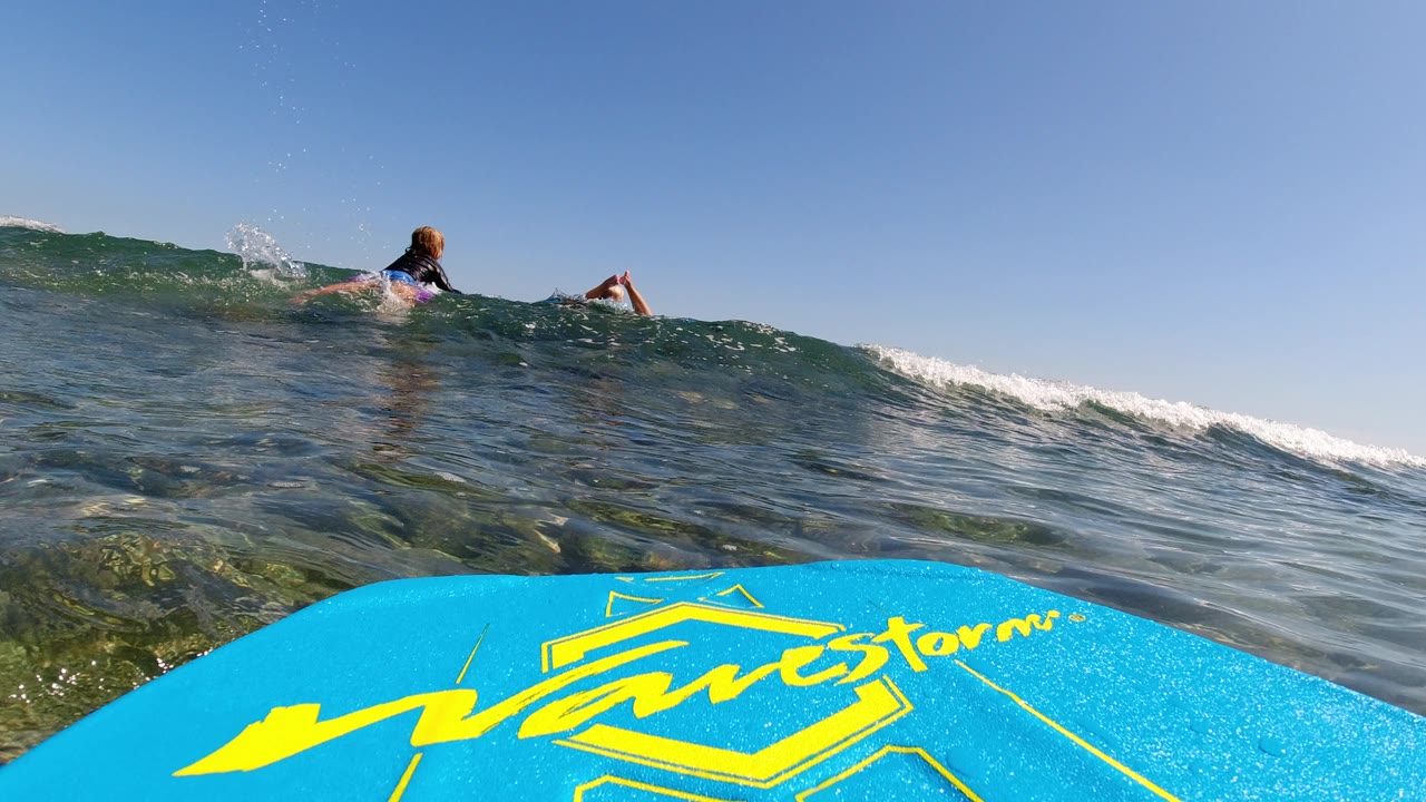 The blue board stays in the foreground while a low wave rises ahead in Los Cabos, Mexico, and small figures remain visible near the wave line.