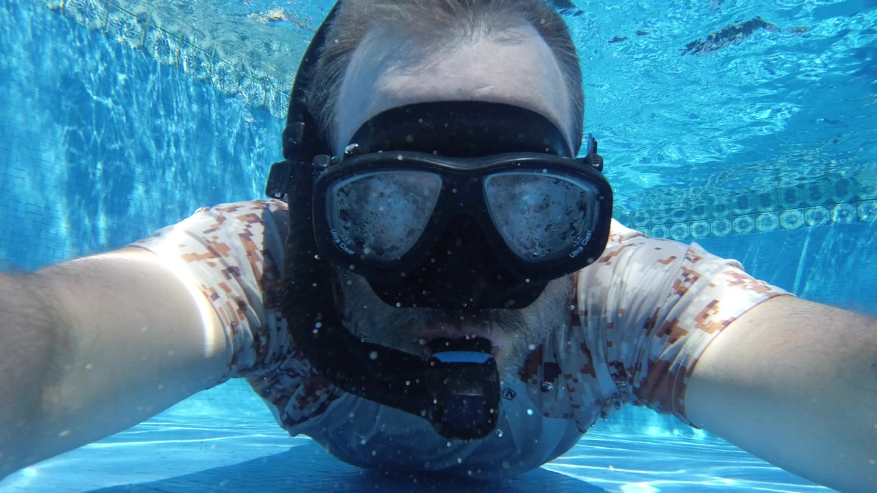 A close underwater frame in Los Cabos, Mexico, shows the swimmer centered with arms forward, black mask over the face, a mouthpiece below, and rippled blue pool water across the background.