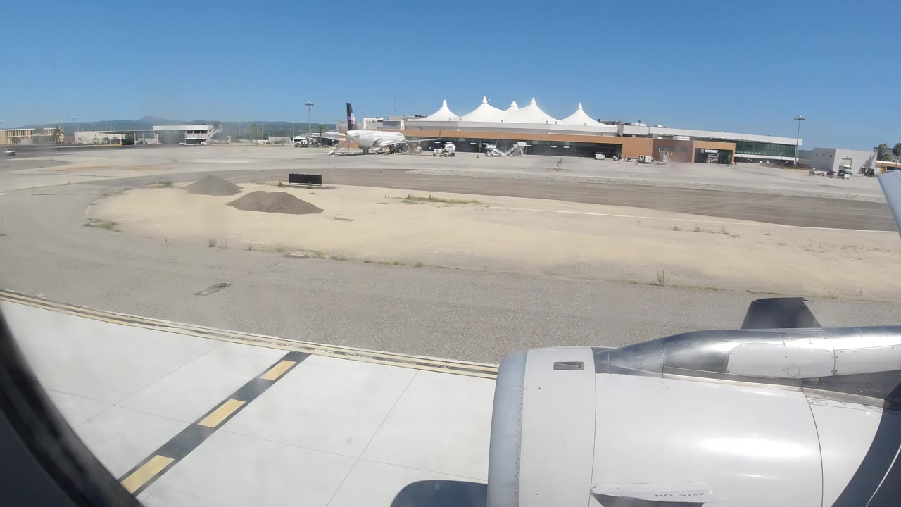 Passenger-window view of a jet engine beside a sunlit taxiway.