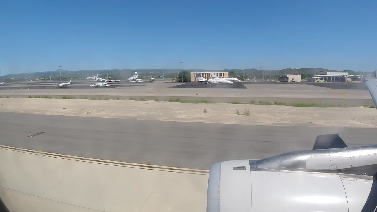 Opening frame from inside a jet at Los Cabos, Mexico, showing the engine in the lower foreground, taxiway pavement, parked aircraft, airport buildings, distant hills, and no visible people.