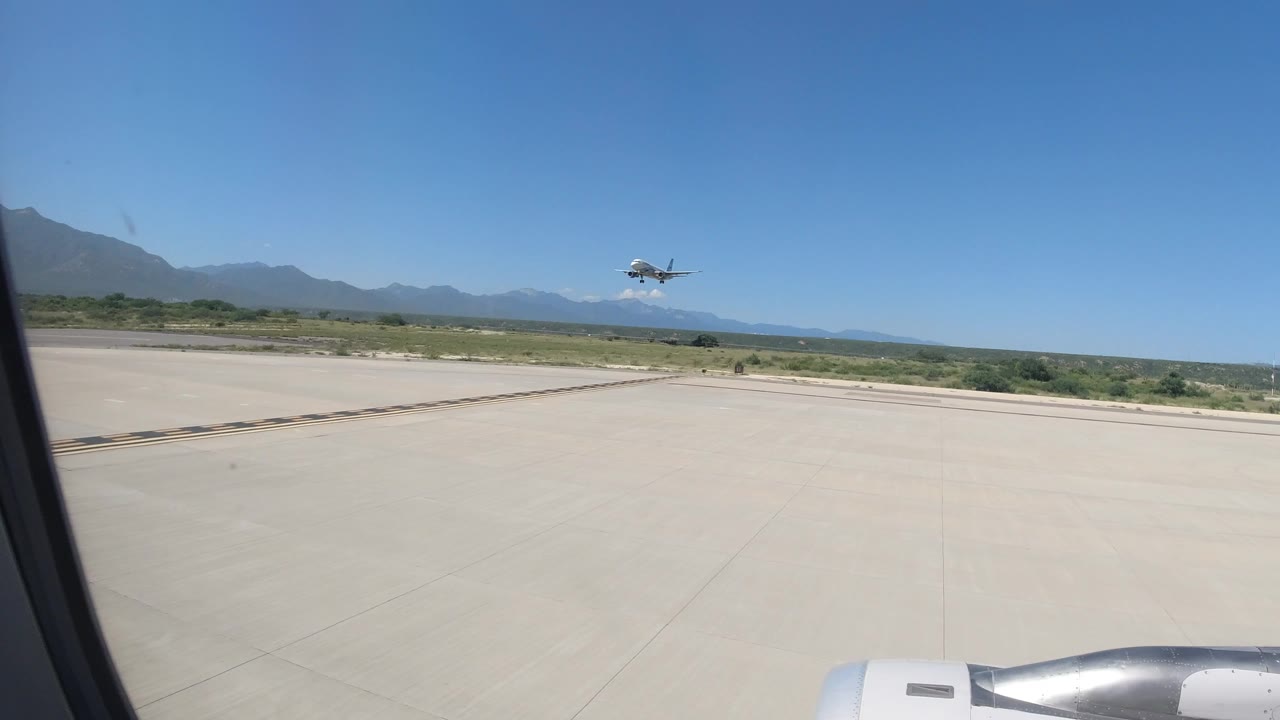At 0:00 in Los Cabos, Mexico, a passenger-window view shows runway pavement, a jet engine edge, distant mountains, and a small aircraft low in the blue sky with no visible people.
