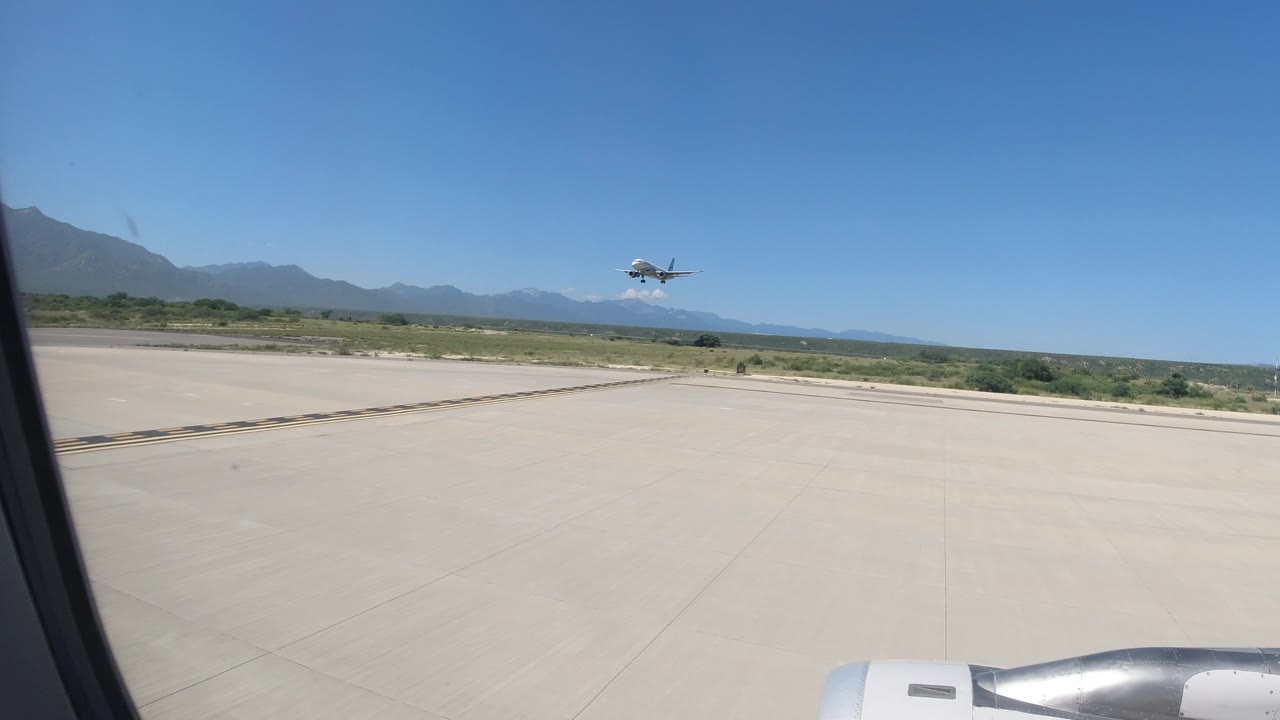 At 0:01 in Los Cabos, Mexico, the distant aircraft appears slightly farther right above the runway, with the window frame and engine still visible and no visible people.