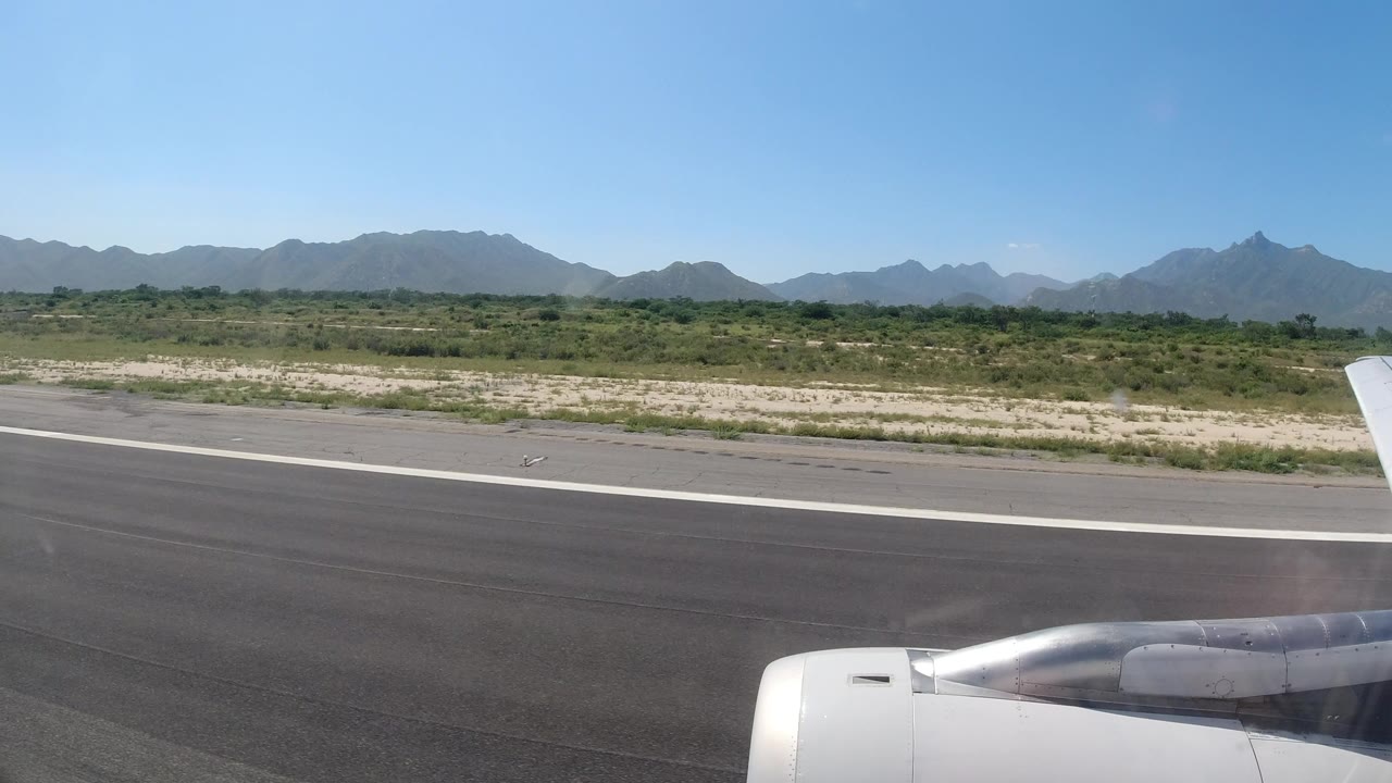 Passenger-window view in Los Cabos, Mexico, showing the airplane engine at the lower right, runway pavement, low green brush, distant mountains, blue sky, and no visible people.