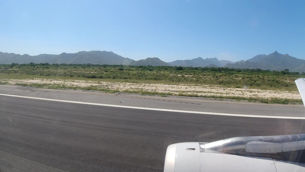 The airplane engine sits along the lower edge as the runway stretches across the frame, with scrubby green terrain and a hazy Los Cabos mountain ridge under clear sky, no visible people.