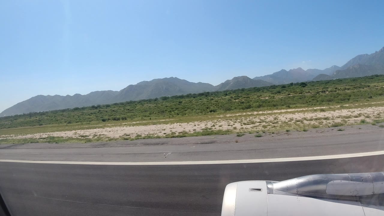 Passenger-window view in Los Cabos, Mexico with an airplane engine in the lower frame, runway pavement, low green scrub, distant mountains, blue sky, and no visible people.