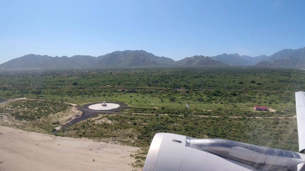 Airplane wing and engine over green fields near Los Cabos, Mexico, with a circular paved area below, mountains on the horizon, and no visible people.