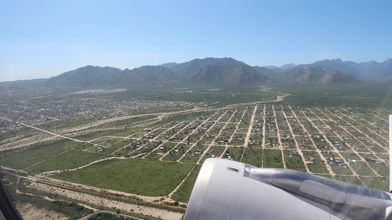 Passenger-window view over Los Cabos, Mexico, with an aircraft wing in the foreground, roads and neighborhoods below, mountains on the horizon, and no visible people.