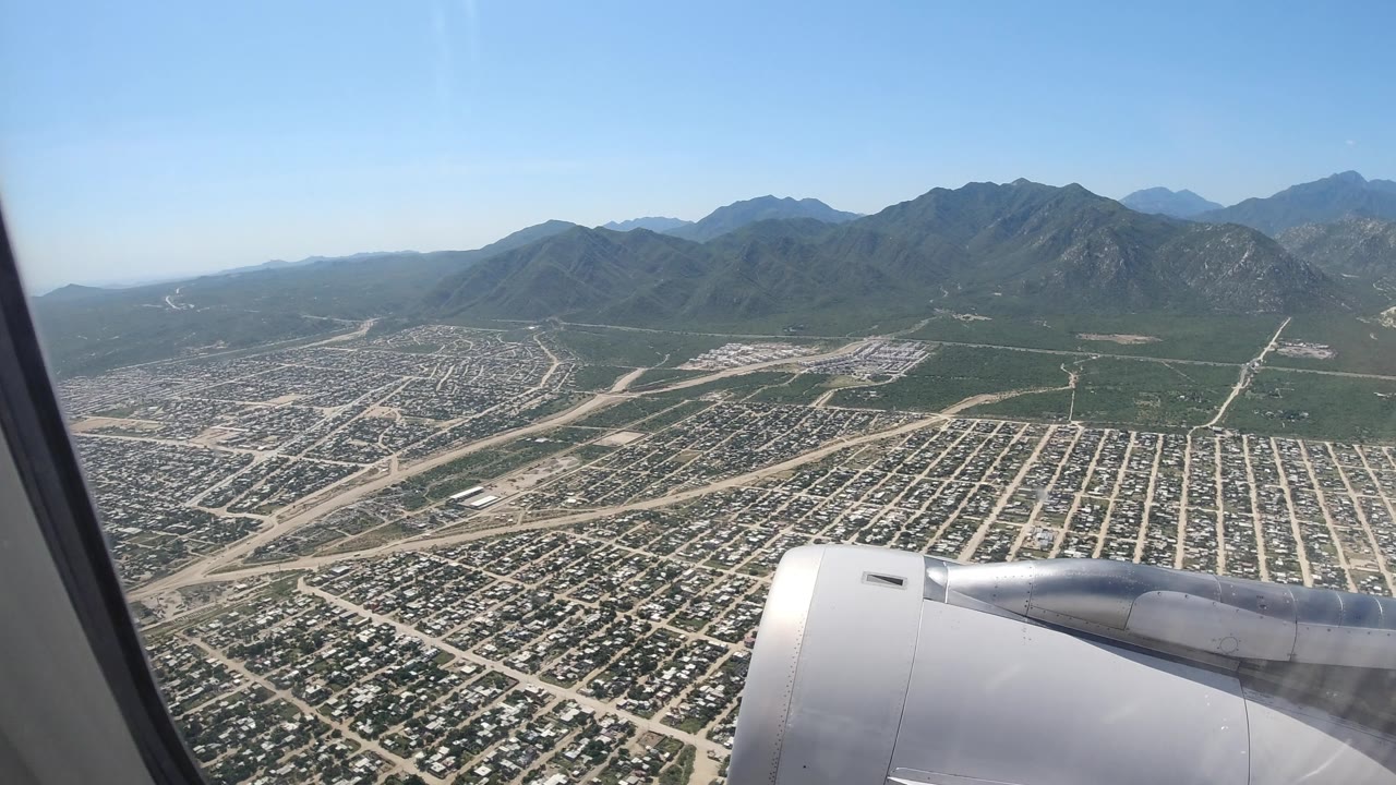 Los Cabos aerial frame with the jet engine lower center, a dense neighborhood grid below, pale roads crossing open terrain, mountain ridges in the distance, and no visible people.