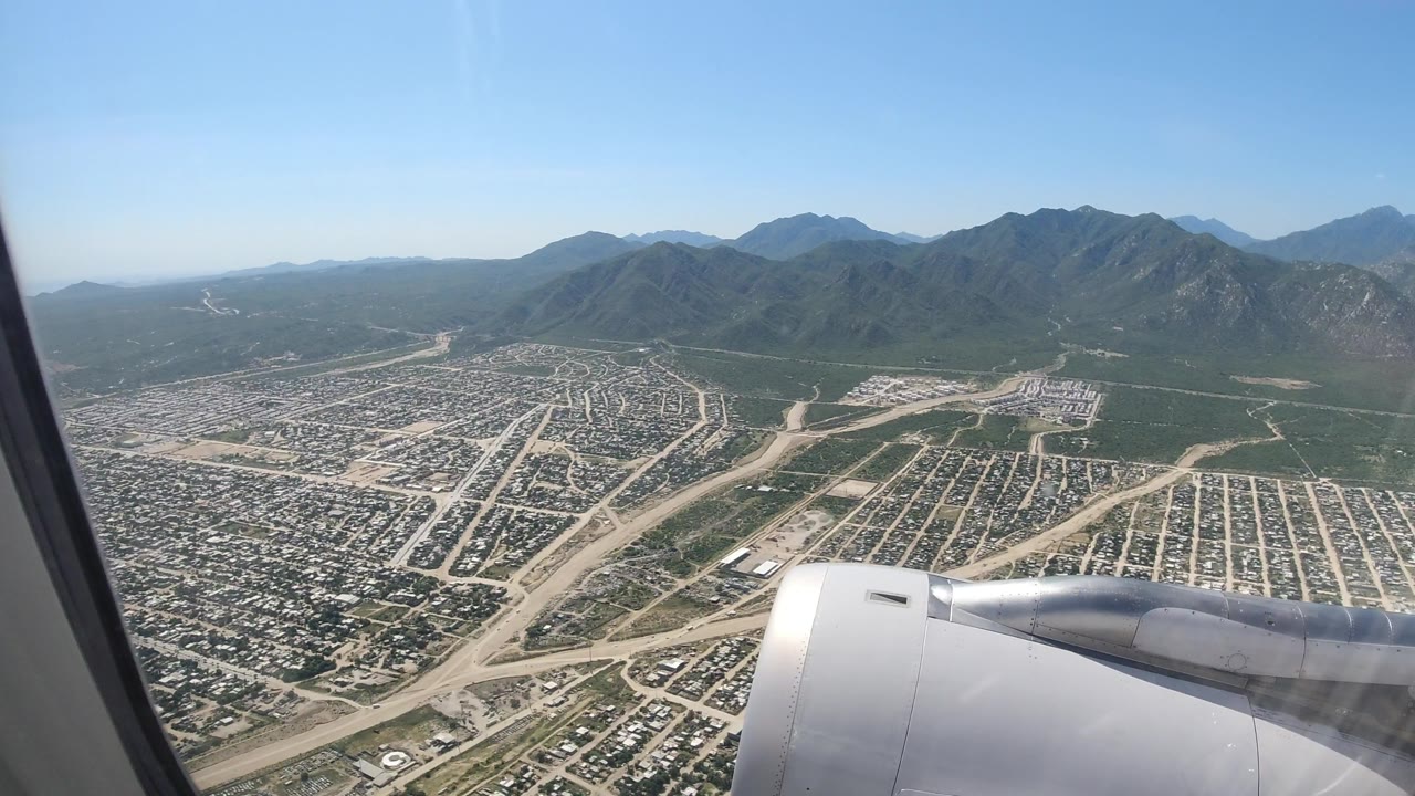 From the airplane window, the engine remains in the lower foreground while neighborhoods, pale road cuts, and mountain ridges fill the Los Cabos, Mexico view with no visible people.