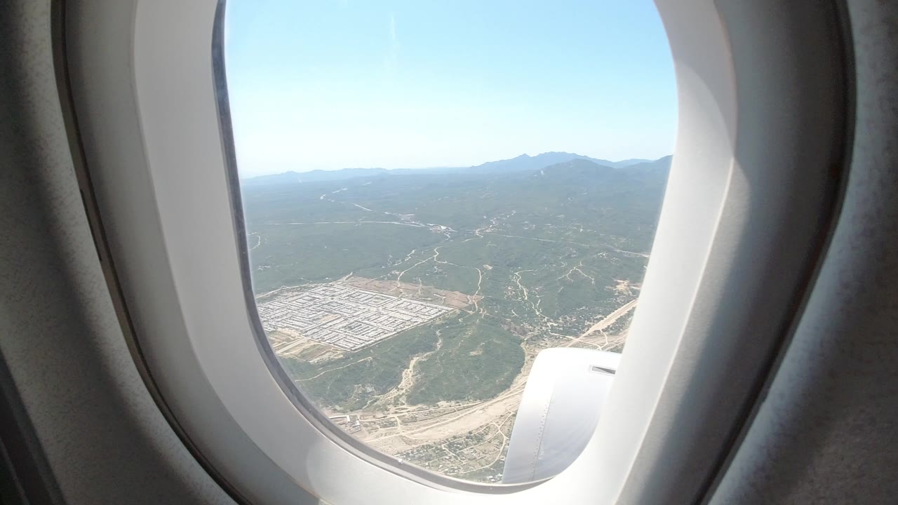 Airplane passenger window view over Los Cabos, Mexico, with green hills, pale roads, a gridded development area at lower left, distant mountains, blue sky, and no visible people.