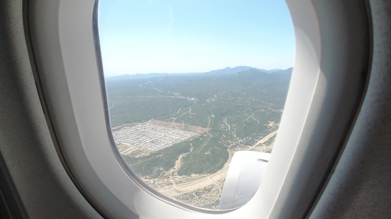 Airplane window frame looking out over Los Cabos, Mexico, with the ground slightly lower in frame, road cuts across green terrain, distant mountains on the horizon, and no visible people.