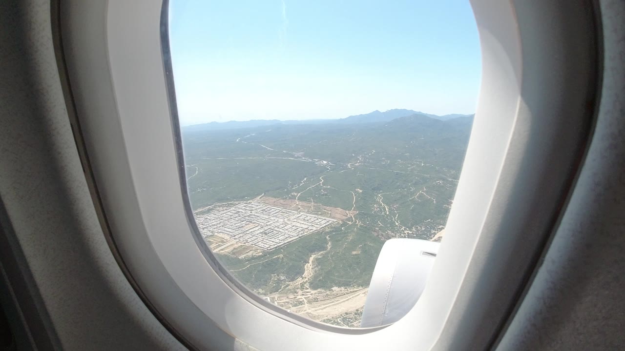 Passenger window view with bright sky, hills, and development below.