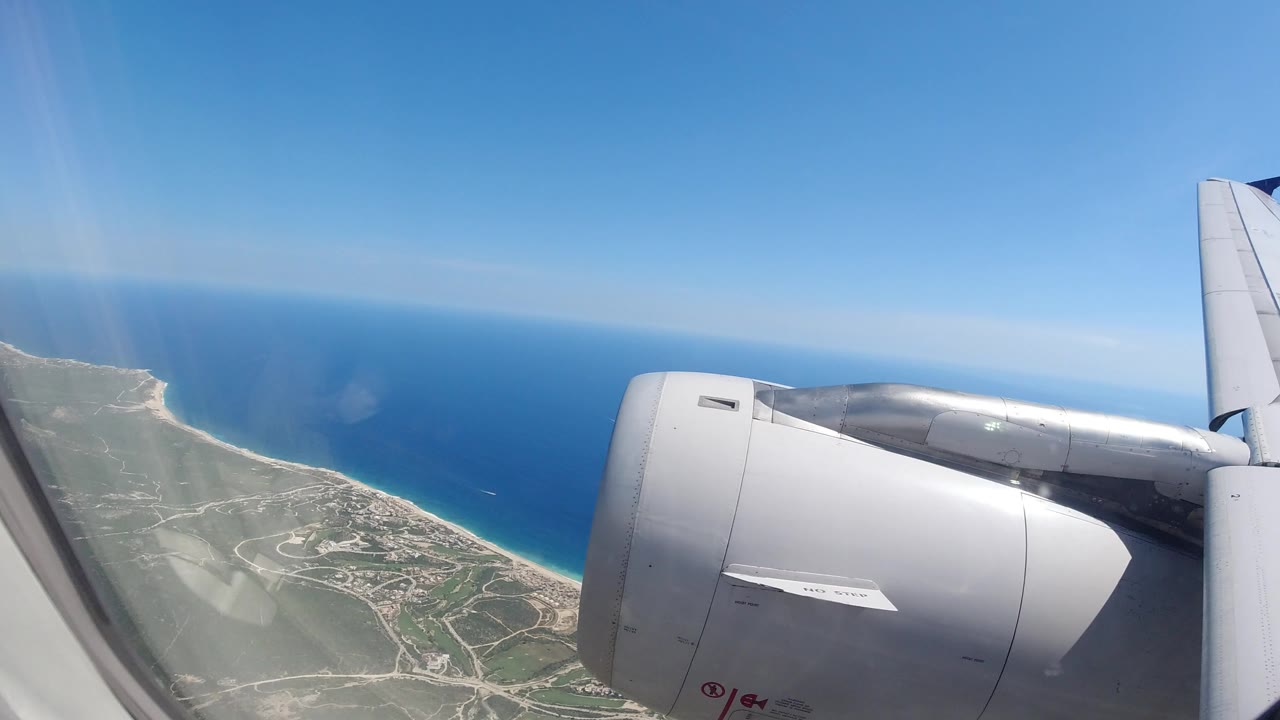 Passenger-window view over the Los Cabos coast with a gray airplane engine and wing in the foreground, blue sea beyond the shoreline, and no visible people.