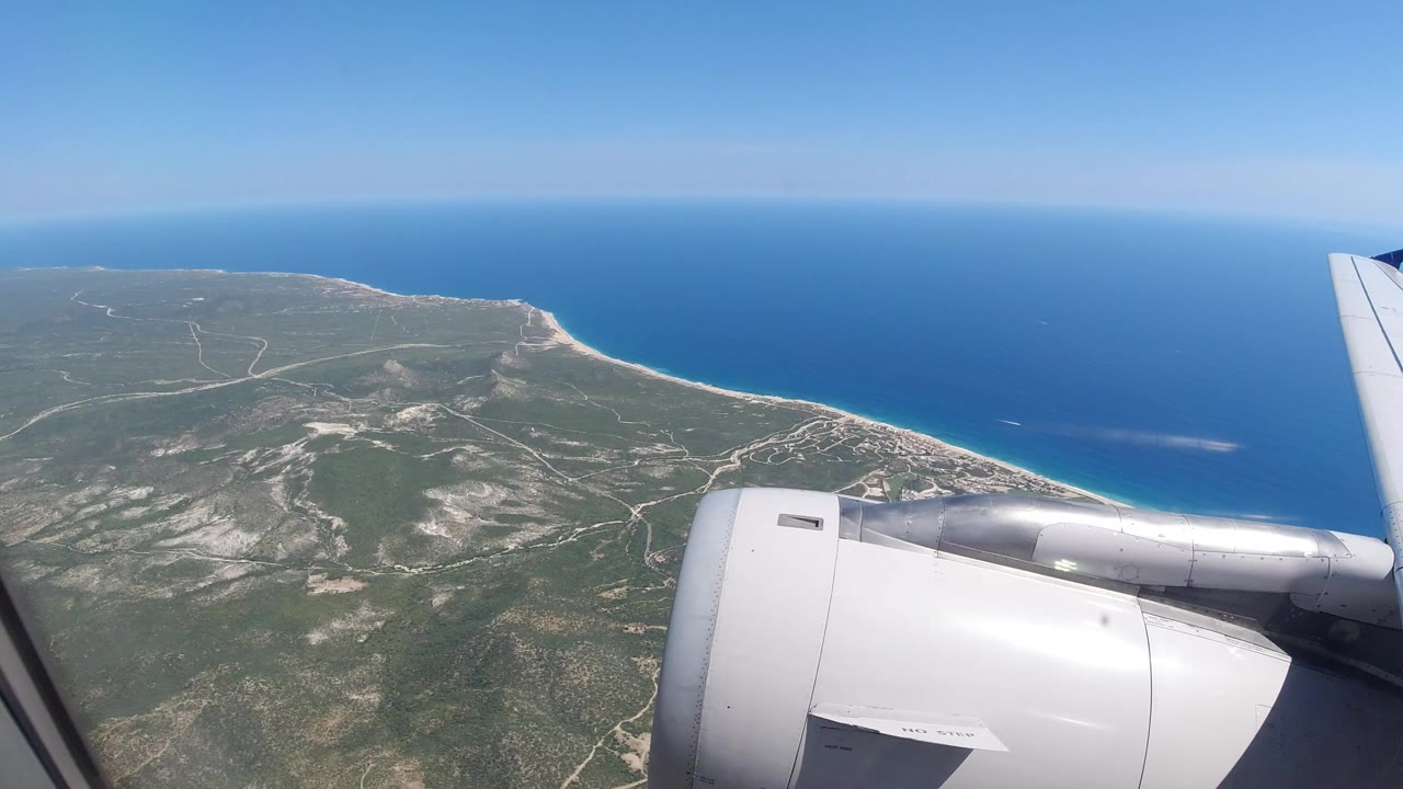 Early view from an airplane window over Los Cabos, Mexico, with a white engine in the lower frame, a wing at right, green coastline below, blue sea beyond, and no visible people.