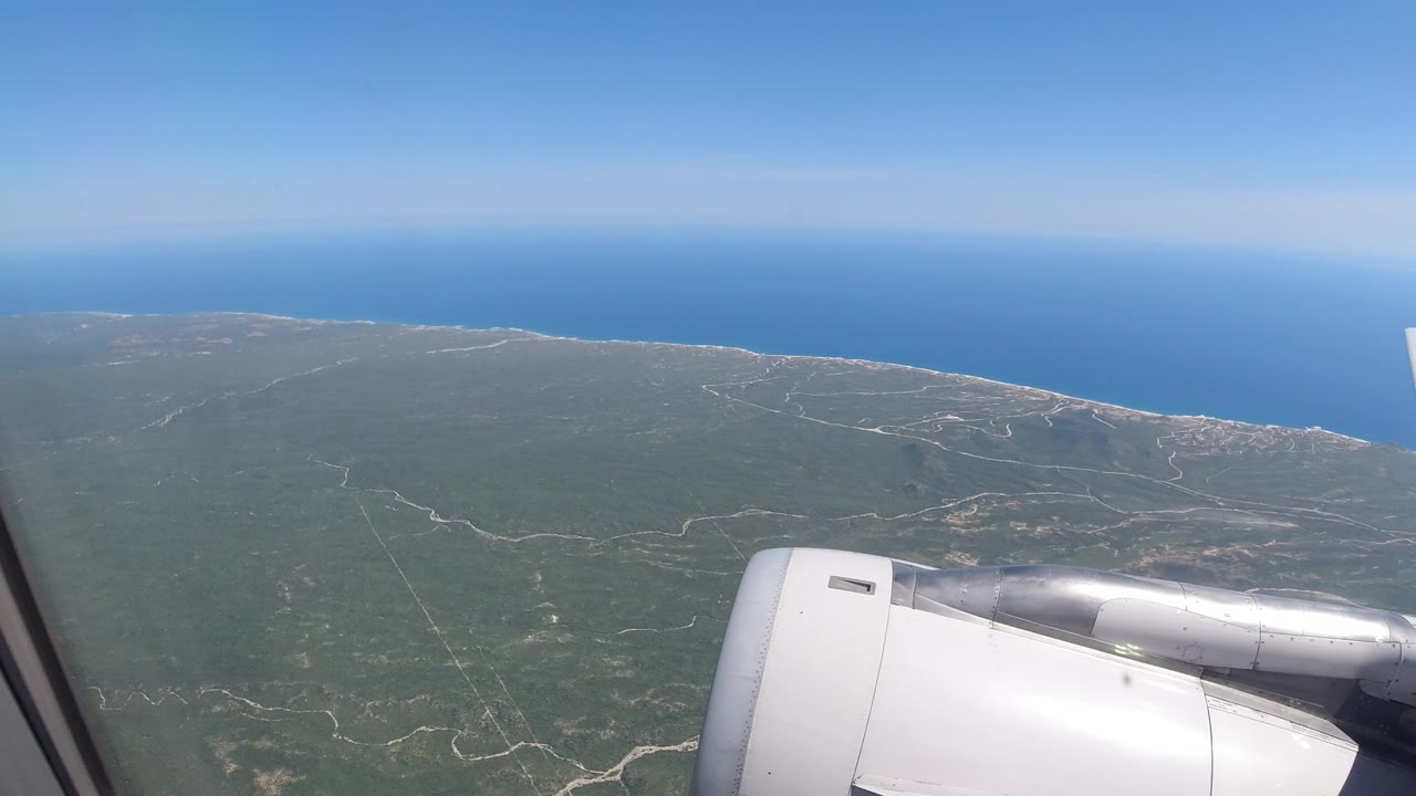 Passenger-window view over Los Cabos, Mexico, with a white airplane engine in the lower frame, green coastal terrain below, blue ocean beyond, and no visible people.