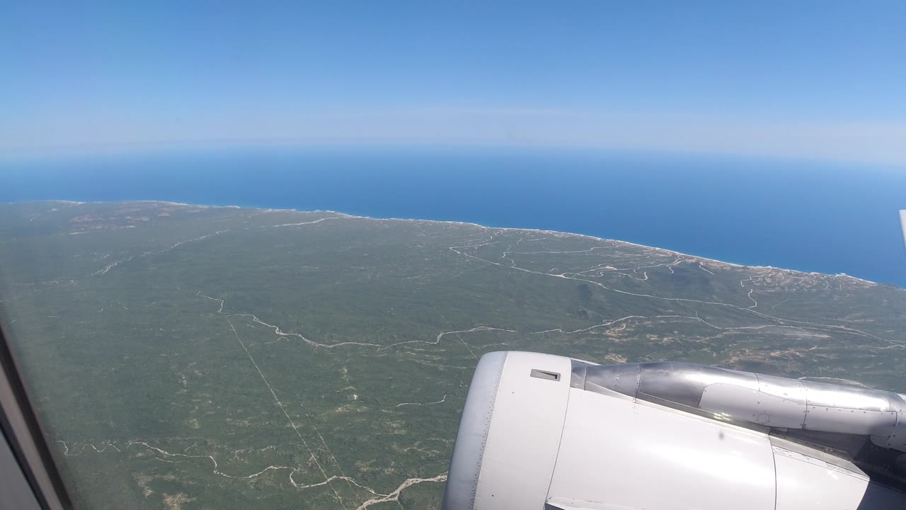 Los Cabos coastline seen from a passenger airplane, with the engine lower right, winding pale roads across green land, a thin shore, blue ocean, and no visible people.