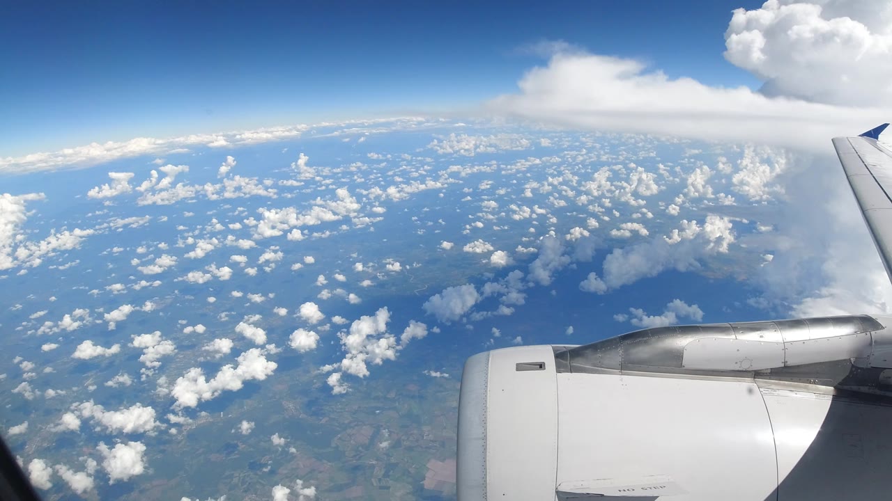 Opening frame of a commercial airplane wing and white engine over scattered cumulus clouds near Los Cabos, Mexico, with blue sky and no visible people.