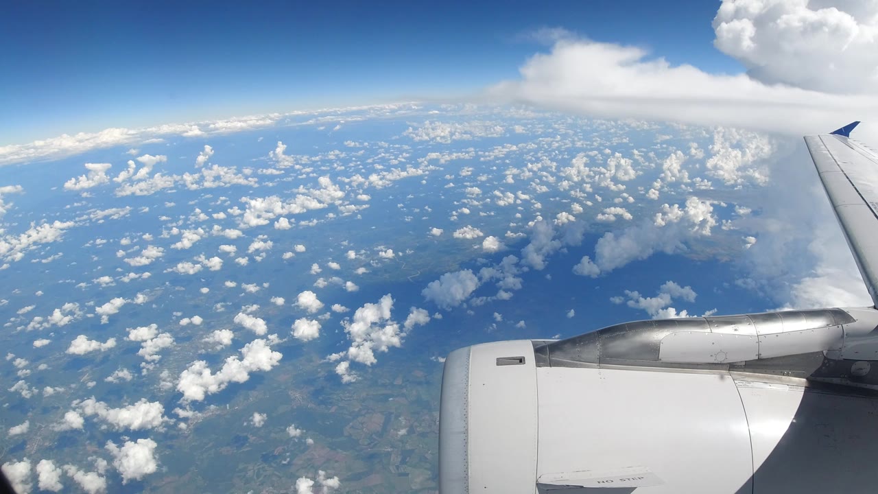 Airplane wing and engine seen from a window over Los Cabos, Mexico, with scattered white clouds, blue sky, distant land and water, and no visible people.