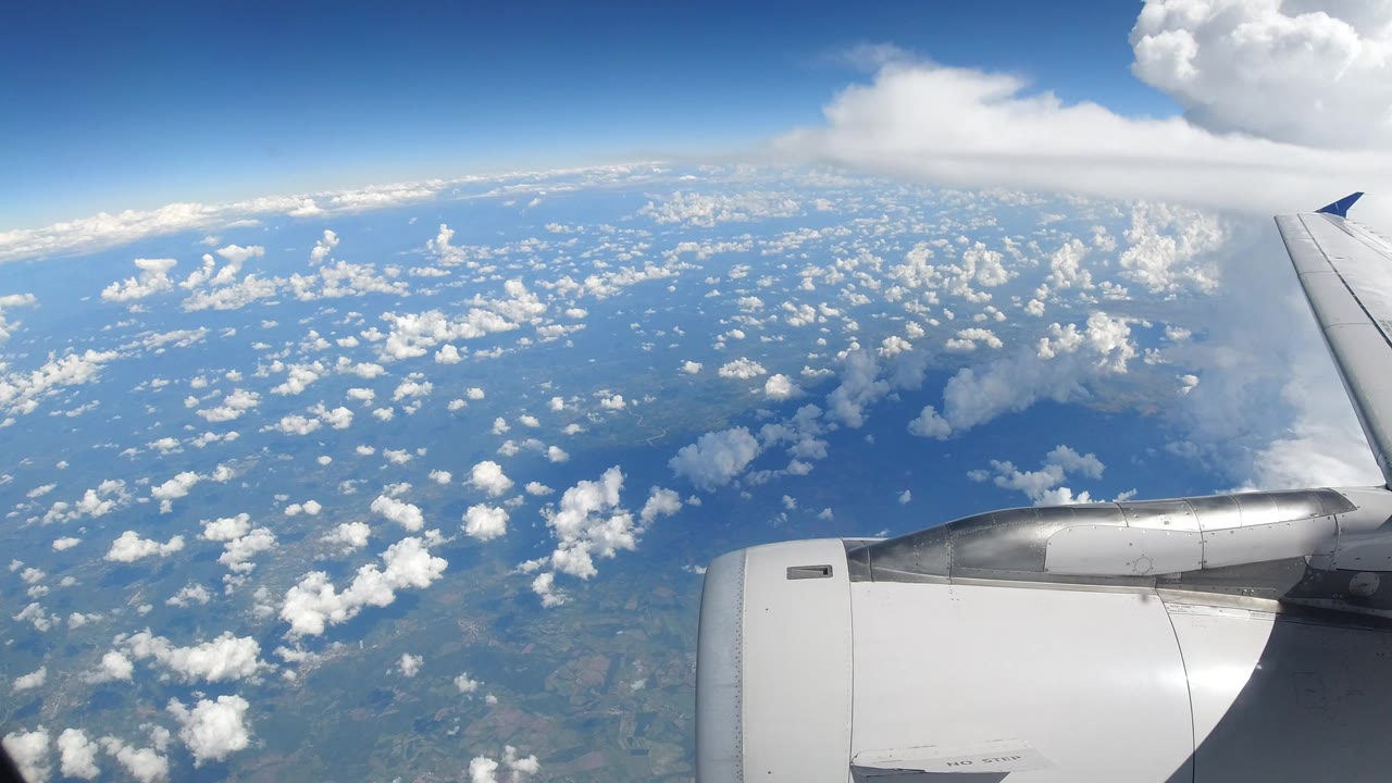 Passenger-plane wing and engine over Los Cabos, Mexico, with a bright cloud field below, a blue upper sky, distant shoreline shapes, and no visible people.