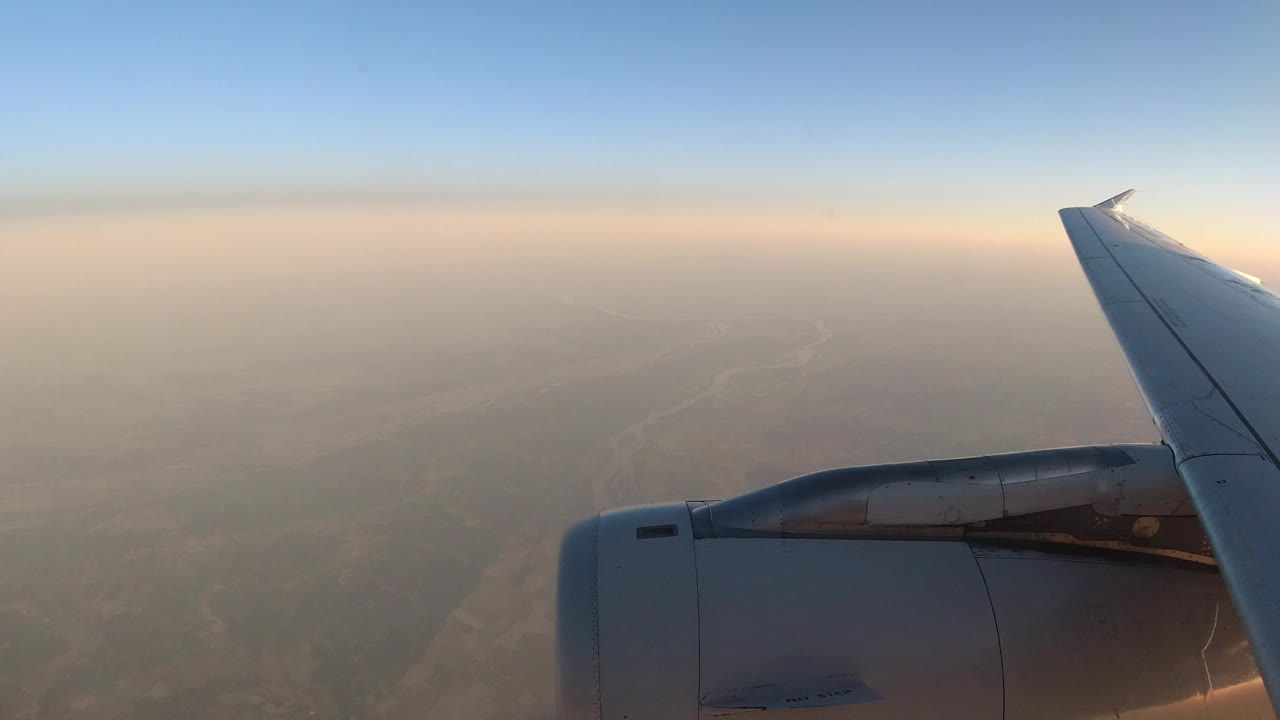 Opening frame from Los Cabos, Mexico footage showing a gray airplane engine and wing against blue sky, a warm hazy horizon, faint land below, and no visible people.