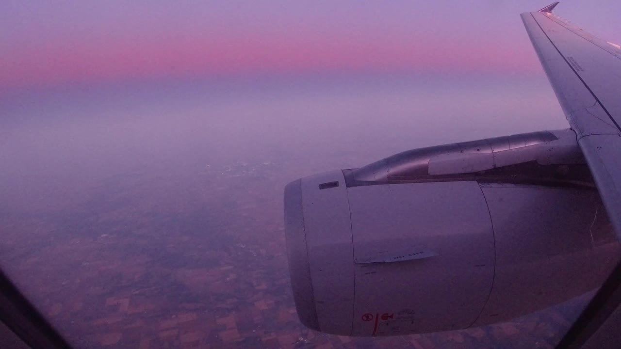 Passenger-window view over Los Cabos, Mexico, with a jet engine and wing in the foreground, hazy land below, a pink-purple horizon, and no visible people.