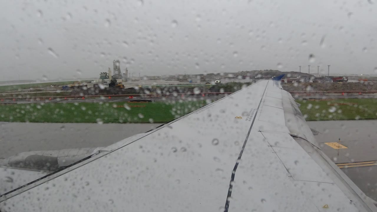 Opening view from an airplane window in Los Cabos, Mexico, with raindrops on the glass, a wing in the foreground, wet runway pavement, distant buildings, and no visible people.