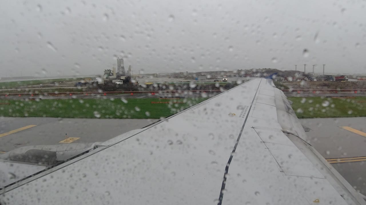 Rainy airplane window looking across a wing toward runway and buildings.