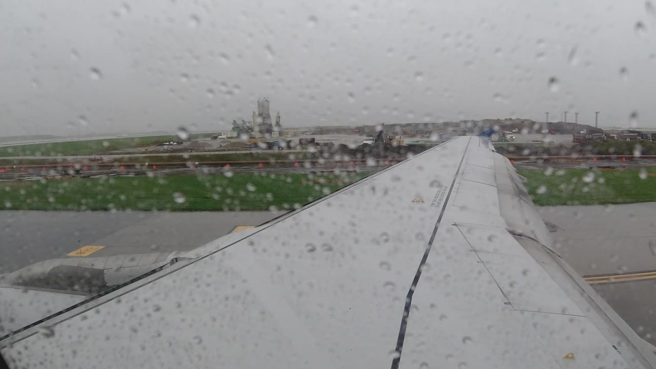 Start of a Los Cabos, Mexico airplane-window view with raindrops on glass, a white wing in the lower frame, wet runway pavement, green grass, gray sky, and no visible people.