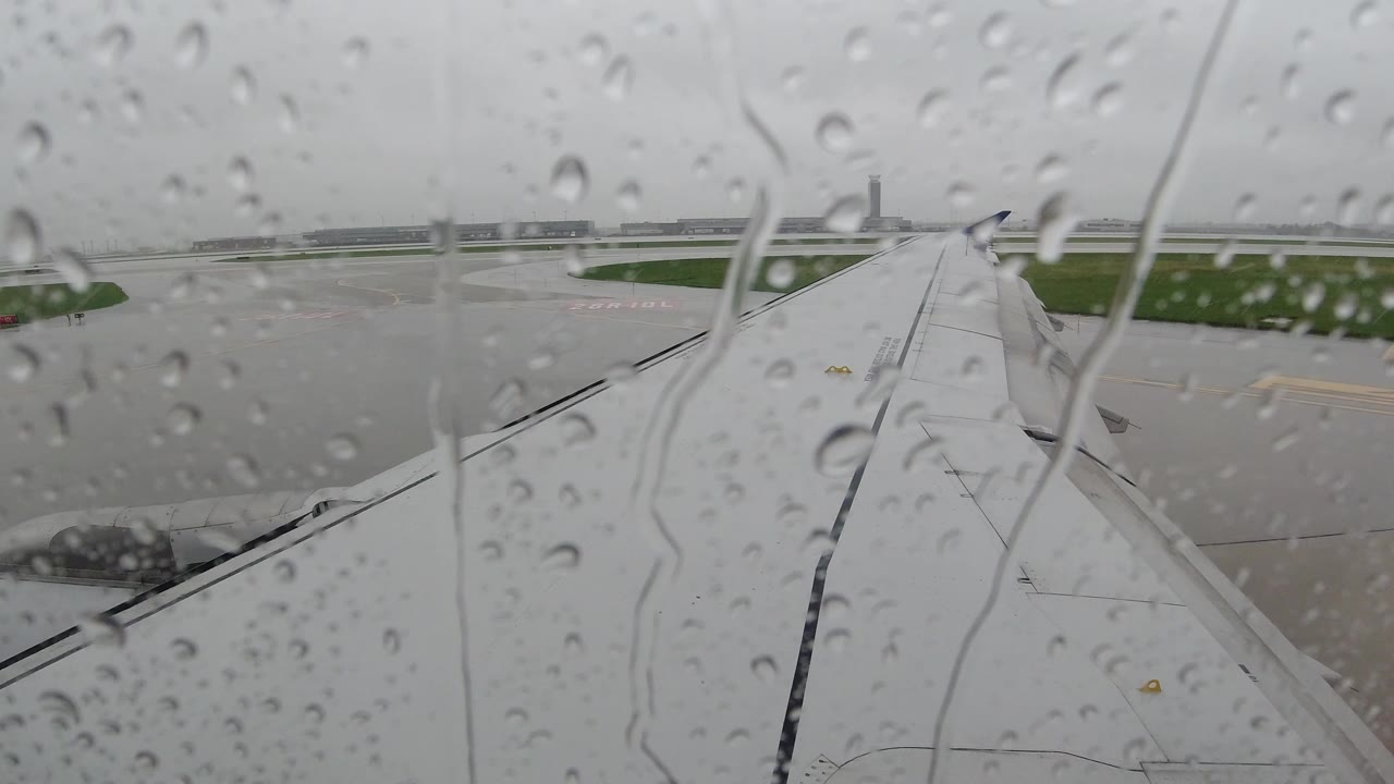 At 0:00 in Los Cabos, Mexico, raindrops cover the aircraft window while a white wing points toward a blurred gray tarmac and low horizon, with no visible people.