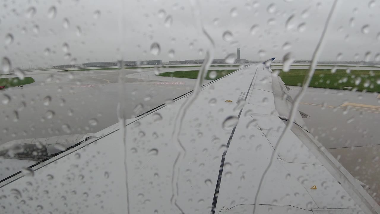 At 0:01 in Los Cabos, Mexico, clustered water beads and vertical streaks sit on the window foreground over a soft view of the airplane wing, tarmac, grass, and cloudy sky, with no visible people.