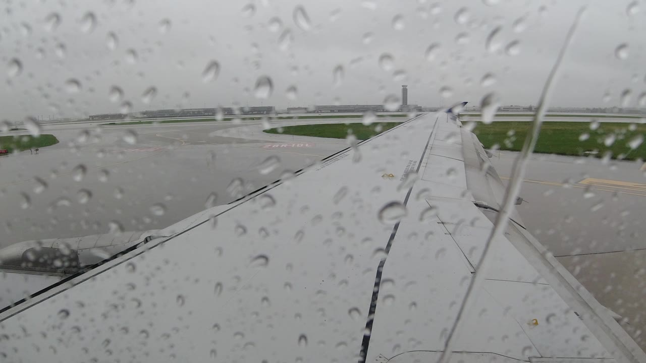 Airplane wing seen through raindrops with airport buildings in the distance.