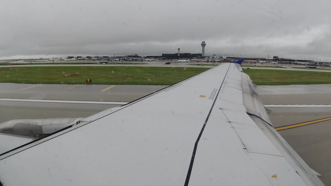 Passenger-window view in Los Cabos, Mexico, with an airplane wing filling the foreground, wet runway pavement, distant airport buildings, a control tower, gray clouds, and no visible people.