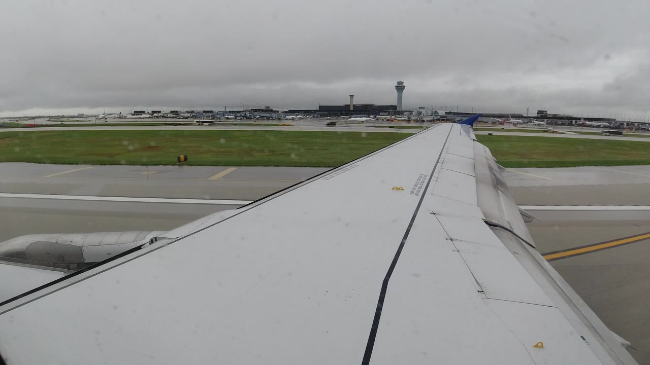 The wing remains centered from the passenger seat as wet pavement, green infield grass, airport buildings, and the control tower sit under low gray clouds in Los Cabos, Mexico, with no visible people.