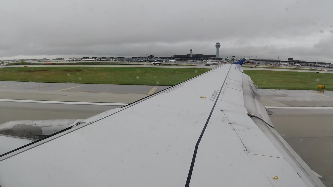 Airplane wing passing airport runway with terminal buildings in the distance.