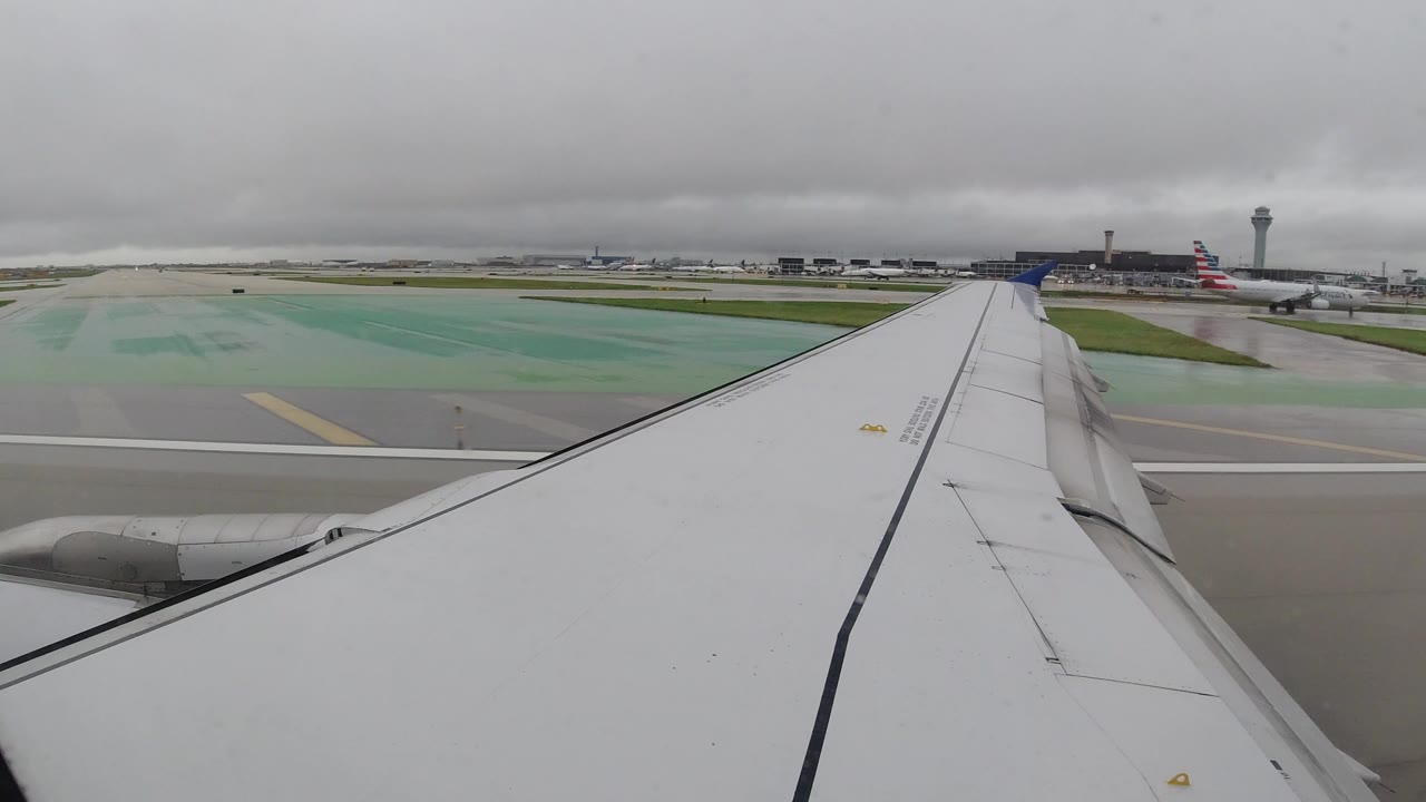 Passenger-window view over a white airplane wing on a wet runway in Los Cabos, Mexico, with airport buildings and a control tower in the distance and no visible people.
