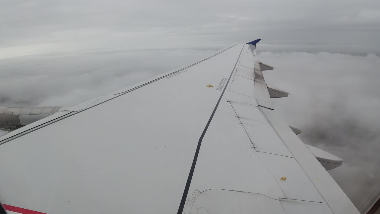 Passenger-window view over Los Cabos, Mexico with a white airplane wing stretching across dense gray-white clouds and no visible people.