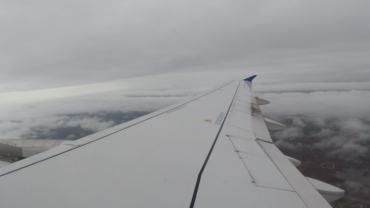 Opening frame of a white airplane wing over broken clouds and dark ground near Los Cabos, Mexico, with no visible people.
