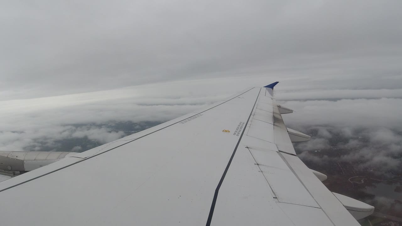 A passenger-window view shows the white wing beneath a low gray cloud layer, with scattered ground visible through clouds over Los Cabos, Mexico, and no visible people.